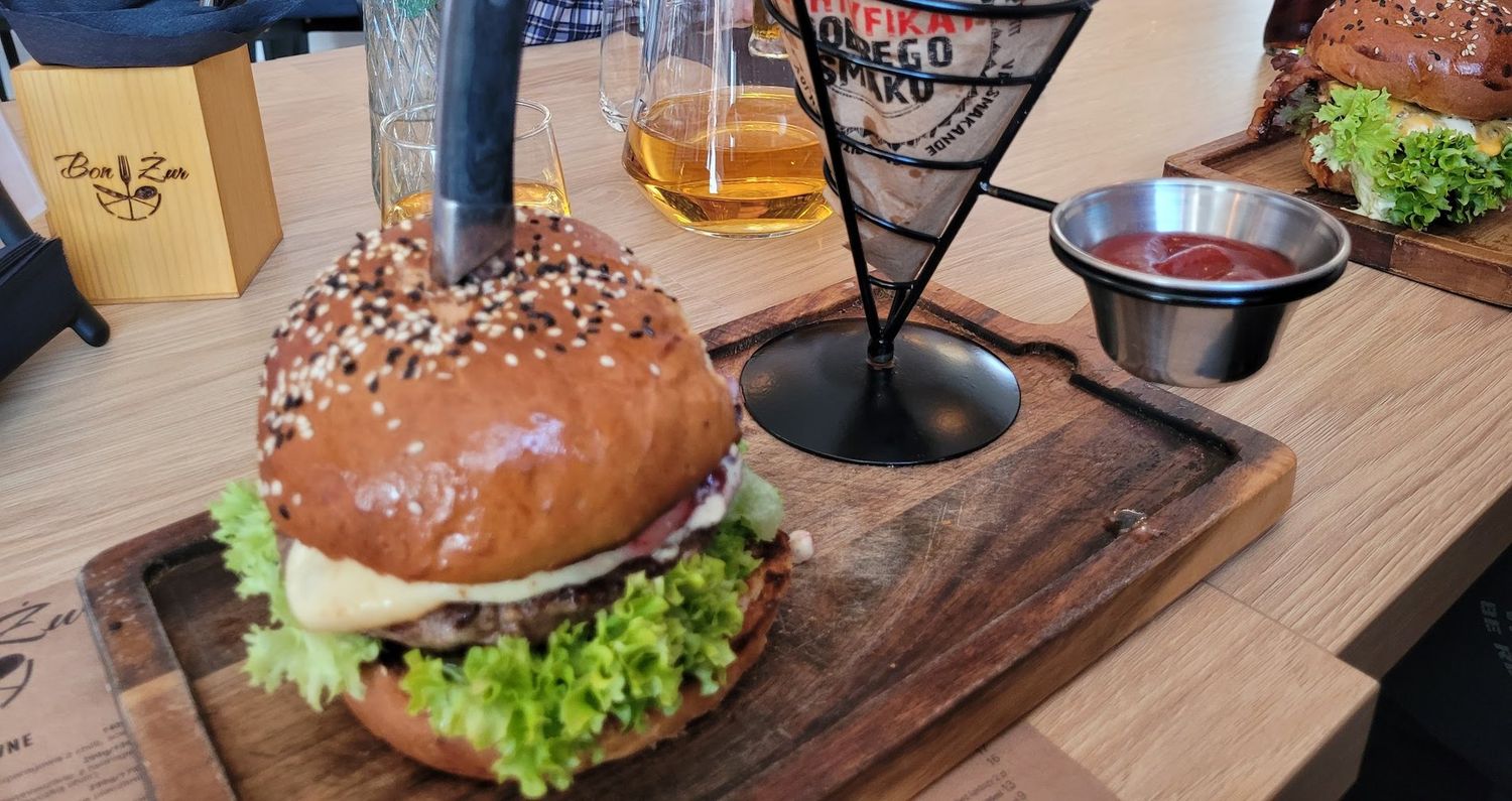 Burger kraftowy z frytkami podany na drewnianej desce obok firmowego pudełka Bon Żur