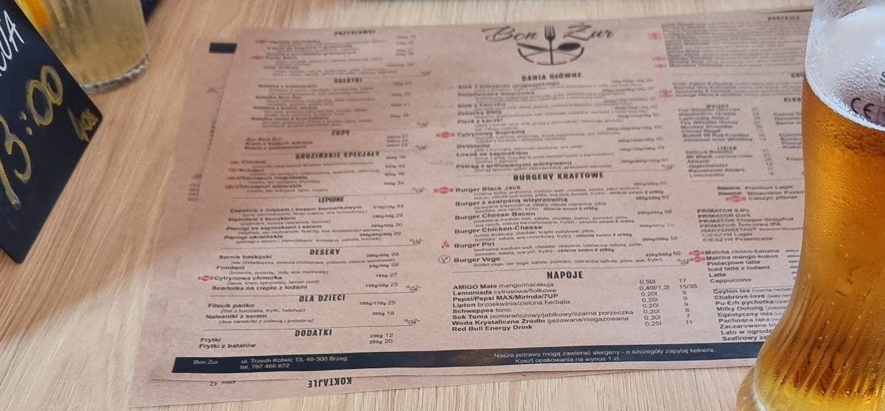Papierowa karta Bon Żur rozłożona na dębowym blacie obok szklanki piwa